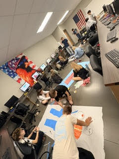 A group of people in a room with computers, working on a large banner on the floor.