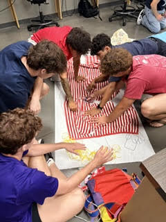 Group of students in a room, collaborating on a large red and white striped fabric spread on the floor.