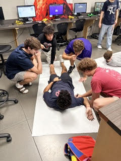 Several people in a room with monitors and computers. One person lies on the floor, another kneels at their feet.
