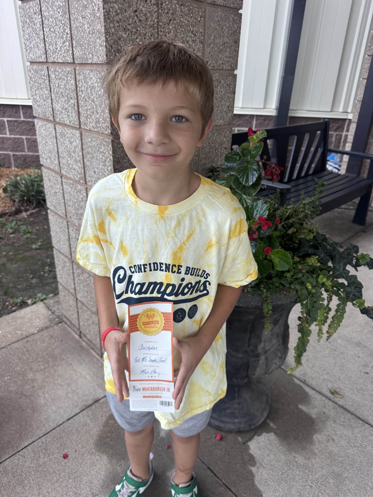 A boy in a yellow shirt holding a certificate stands on a porch next to a potted plant.