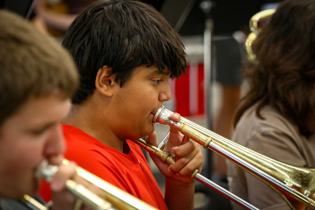 Two boys play trombones. One boy wears a red shirt. A girl with brown hair plays an instrument behind them.