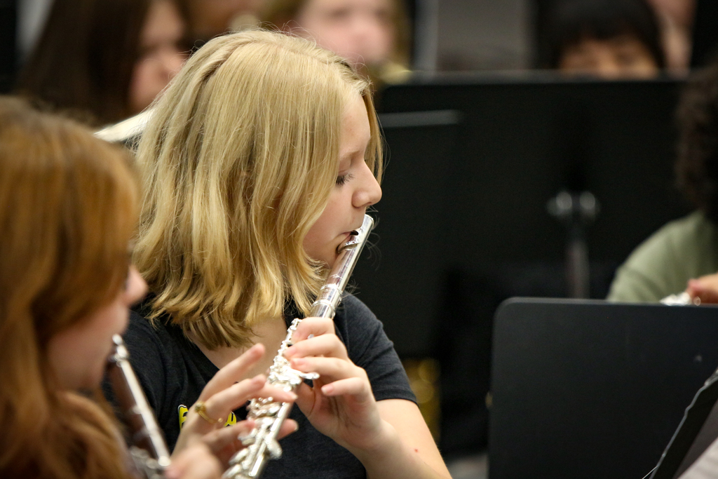 Two girls play flutes in a band, with blurry band members in the background. One girl has blonde hair, and the other has red hair.