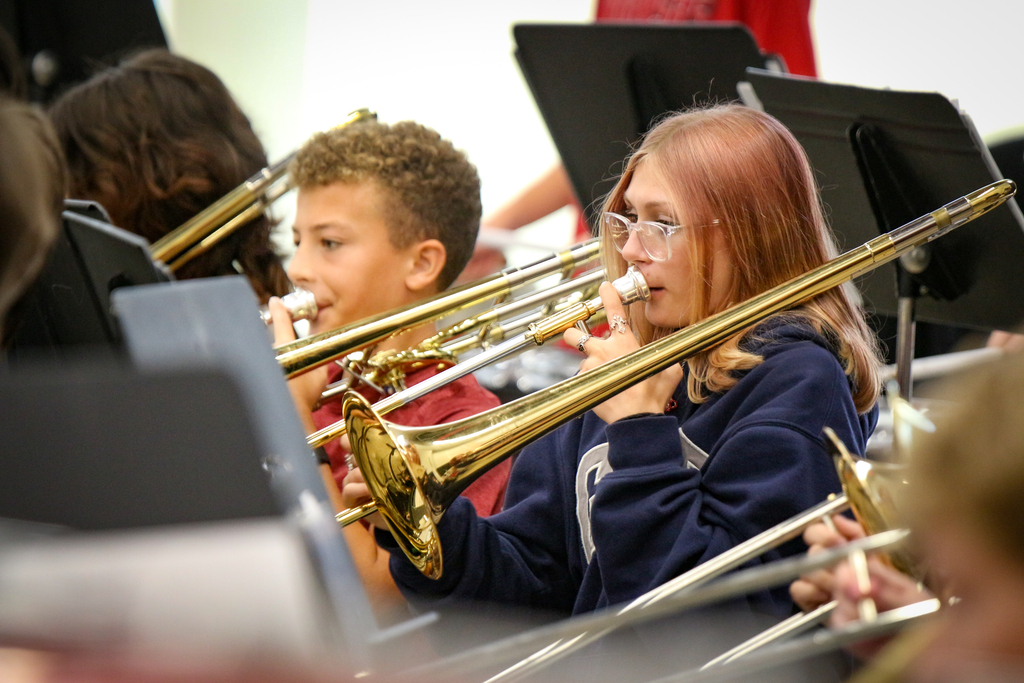 Multiple children in a school band, each holding and playing a trombone. The children are surrounded by other trombone players.