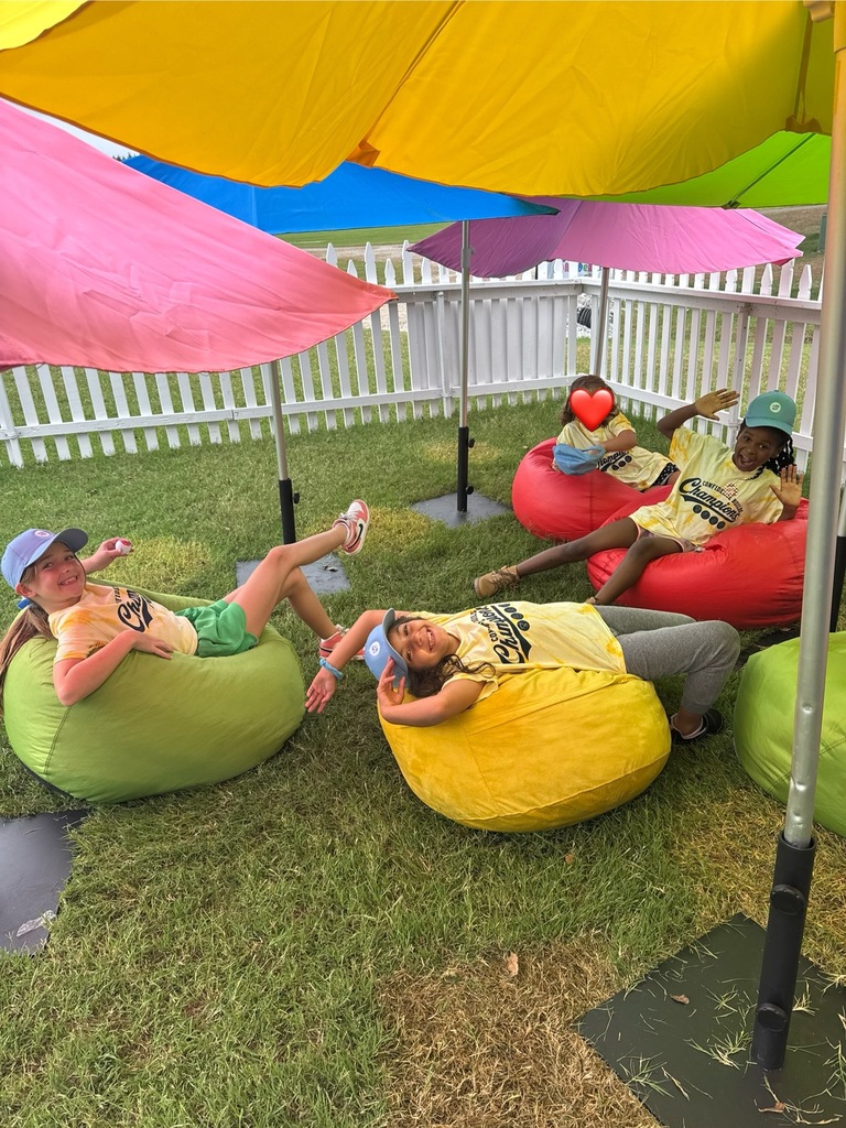 Four children in matching yellow shirts, sitting on bean bags under multicolored umbrellas in a grassy area.