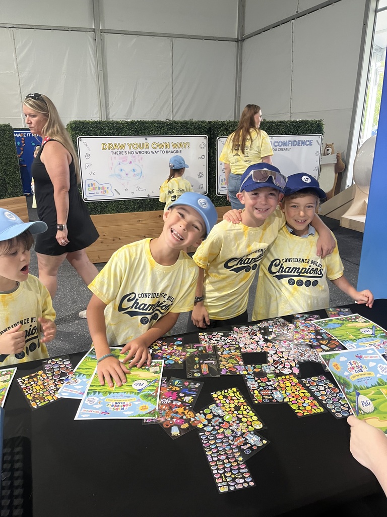 Four kids in yellow shirts and blue caps work on puzzles at a table, with a person in a black dress in the background.