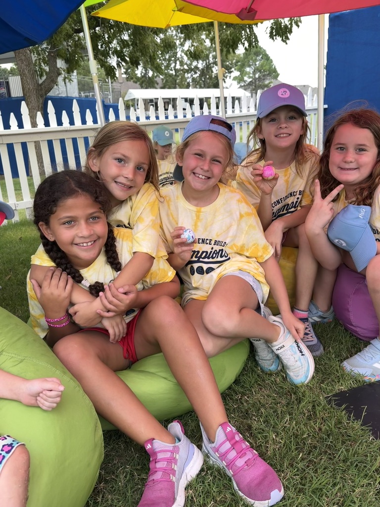 Seven young girls in yellow shirts sit on bean bags on grass. One holds a ball. Umbrellas are above them.