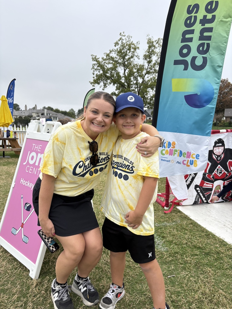 A woman and a child stand on a grass field. Both wear yellow shirts with the word "Champions" on them. Behind them is a colorful banner.