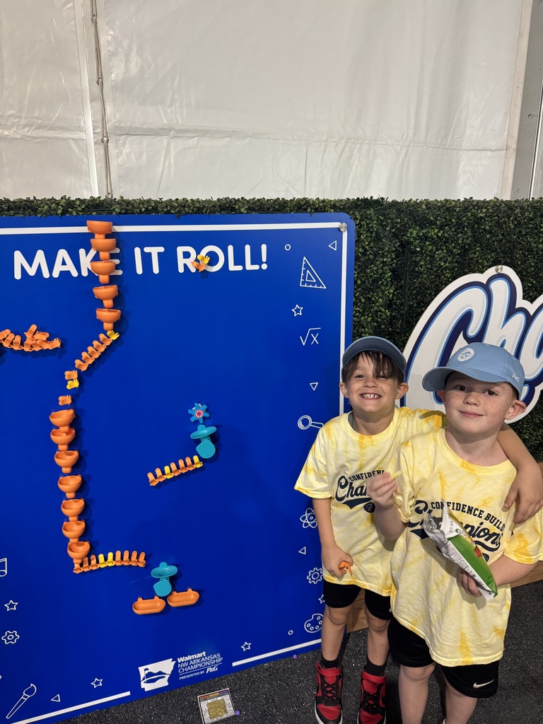 Two young children in matching yellow shirts and hats stand in front of a blue board with "Make It Roll" written on it.
