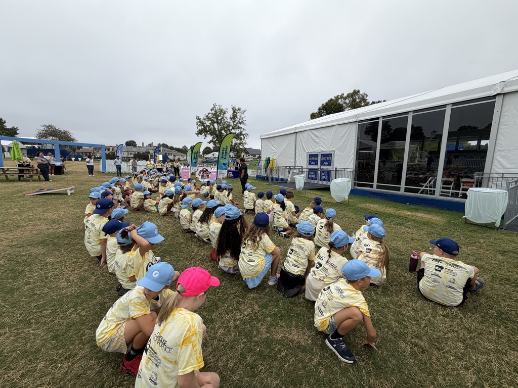 A group of children in yellow shirts and hats sit in a circle on a grassy field with a tent in the background.