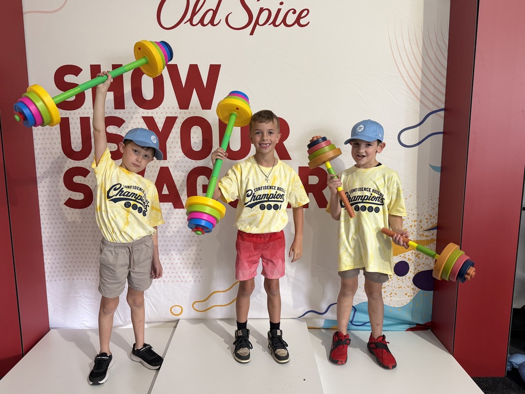Three young boys stand on a white platform, holding colorful dumbbells, with a white wall behind them.