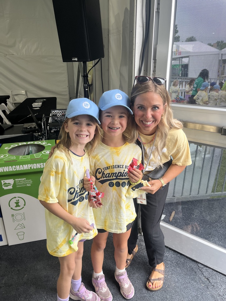 Three people in matching yellow T-shirts smile at the camera. The two girls hold items and the adult wears sunglasses. A tent and speaker are in the background.