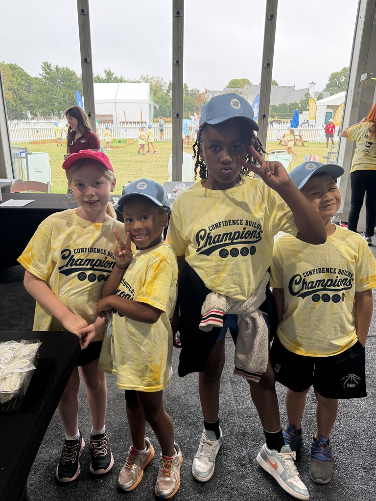 Four children wearing yellow Champion shirts and caps, standing together near a window. A table and trees are visible in the background.