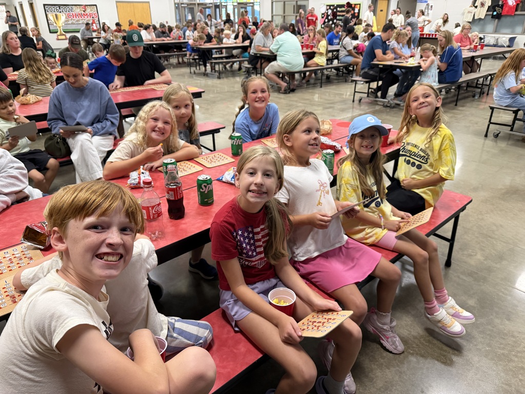 Several children, some with pizza, sit at tables in a school cafeteria. A few others sit at tables in the background.