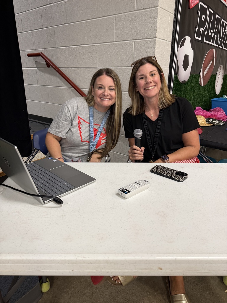 Two women sit at a table in a room with a banner of sports. A laptop, remote, and phone are on the table.