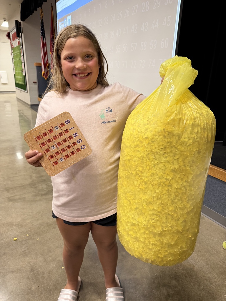 Girl holds bingo card and bag of yellow popcorn in school hallway. Bright yellow bag stands out against gray floor.
