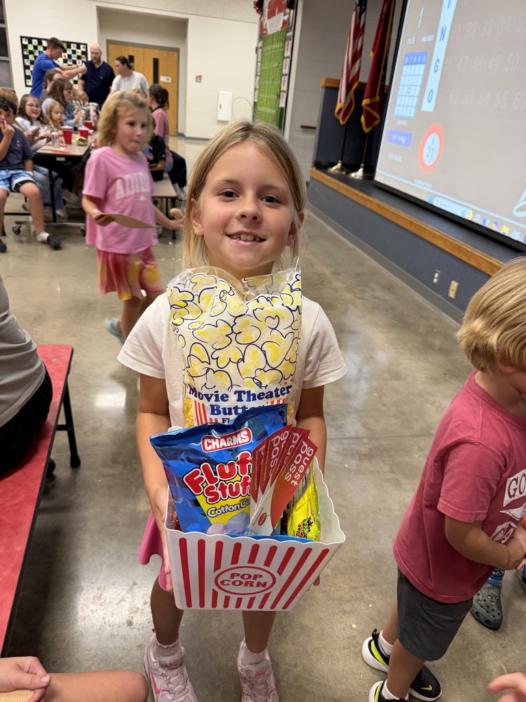 Girl holds a box of snacks while standing in a room with children and a large screen in the background.