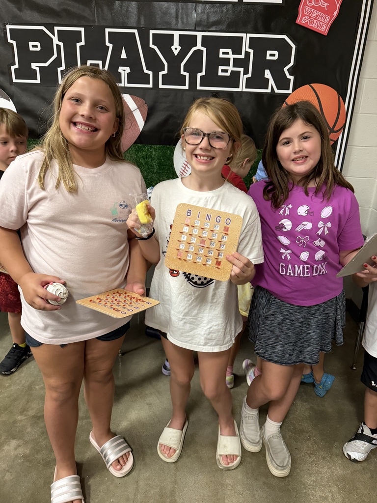 Three girls pose together in front of a sports-themed banner with a basketball and football.