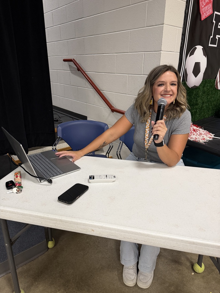 Person at a table with a laptop and phone, holding a microphone. Behind is a black curtain and a soccer poster.