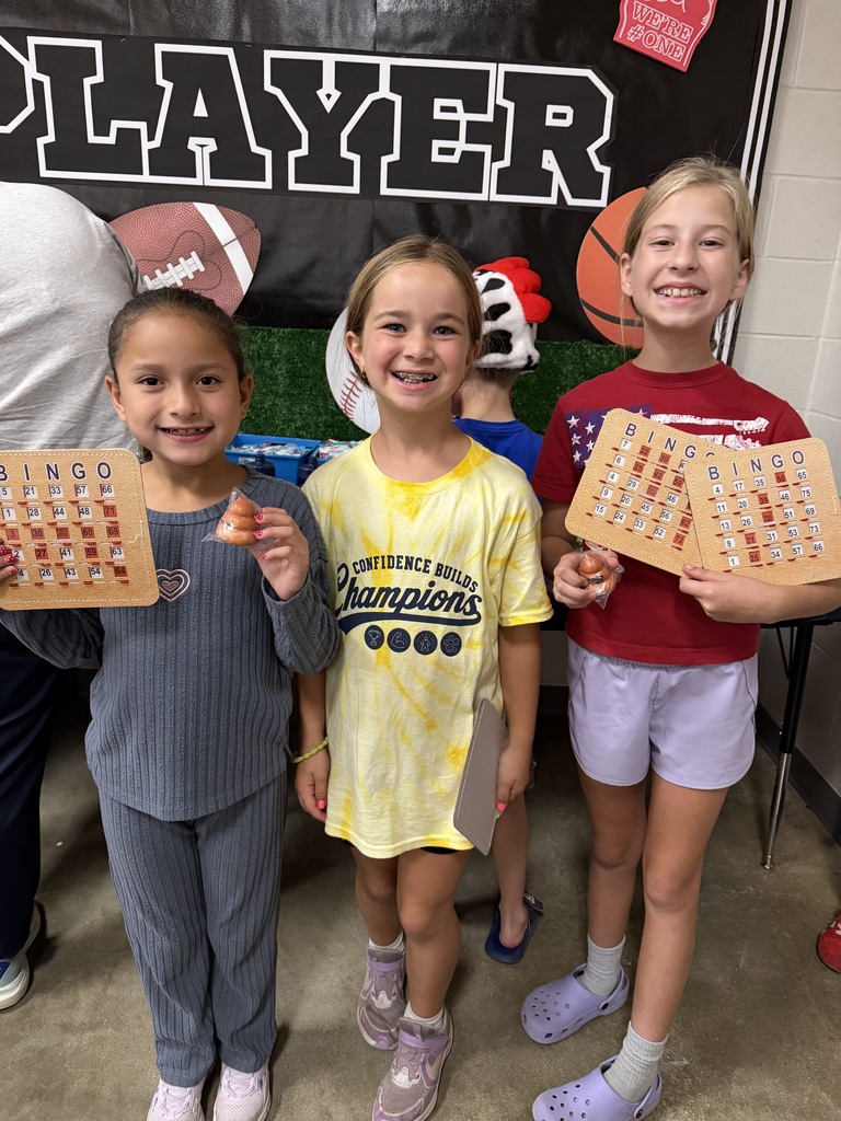 Three young girls smile at camera, holding up boards with puzzles. One wears a yellow shirt, another a red shirt. They stand in front of a banner with sports images.