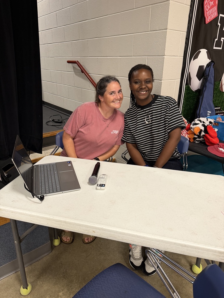 Two women sit at a white table with a laptop, microphone, and other items. Behind them, a wall with a black curtain and sports gear.
