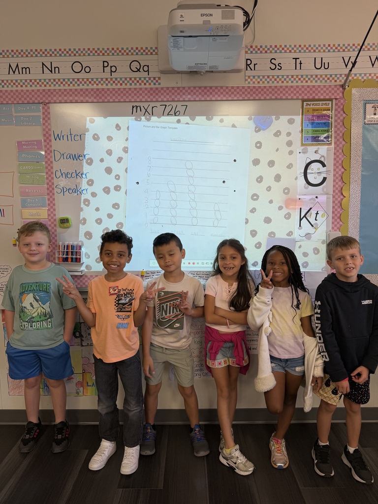 Six children standing next to each other in a classroom. A projector screen is visible in the background.