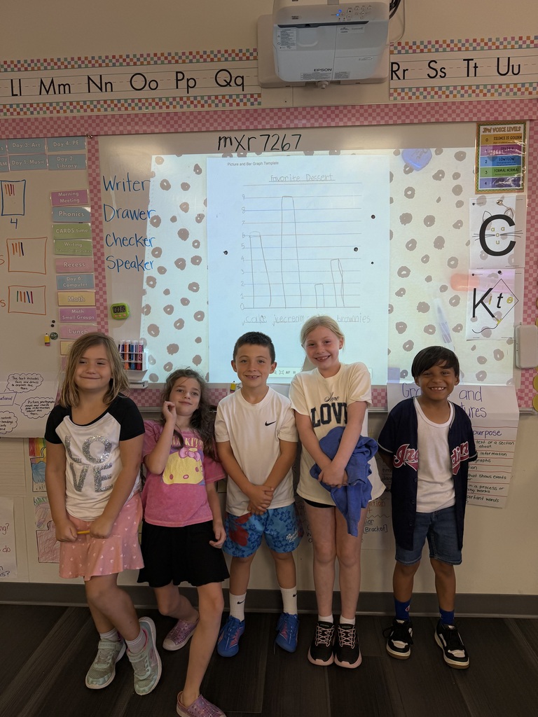 Six children stand before a whiteboard with a projected image, smiling. A bulletin board with alphabet letters is visible.