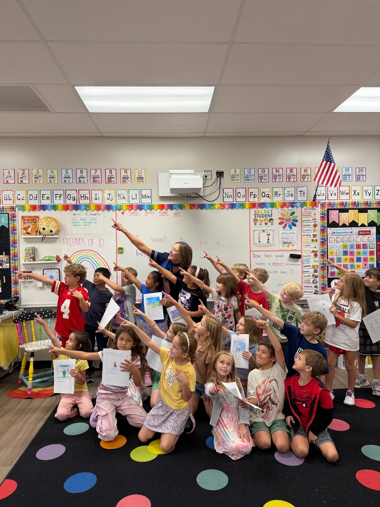 Classroom with kids and teacher. Teacher stands with arms raised. Children sit on floor, hold certificates, and pose for photo. Bright, colorful classroom.