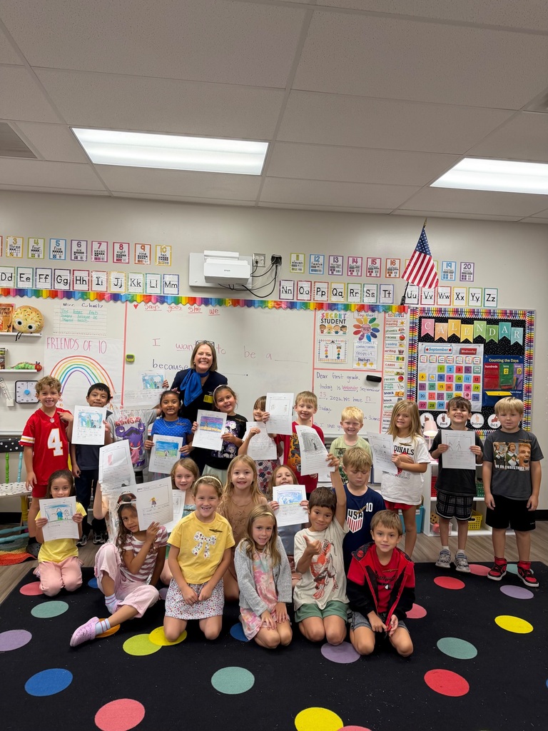 A classroom with a teacher and students holding certificates. A carpet has colorful dots.