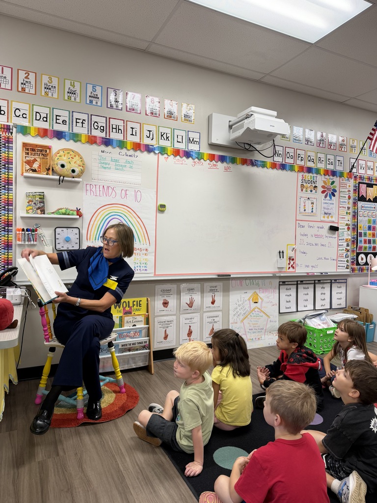 A teacher reads a book to children sitting in a circle. A whiteboard with a rainbow and alphabet is in the background.