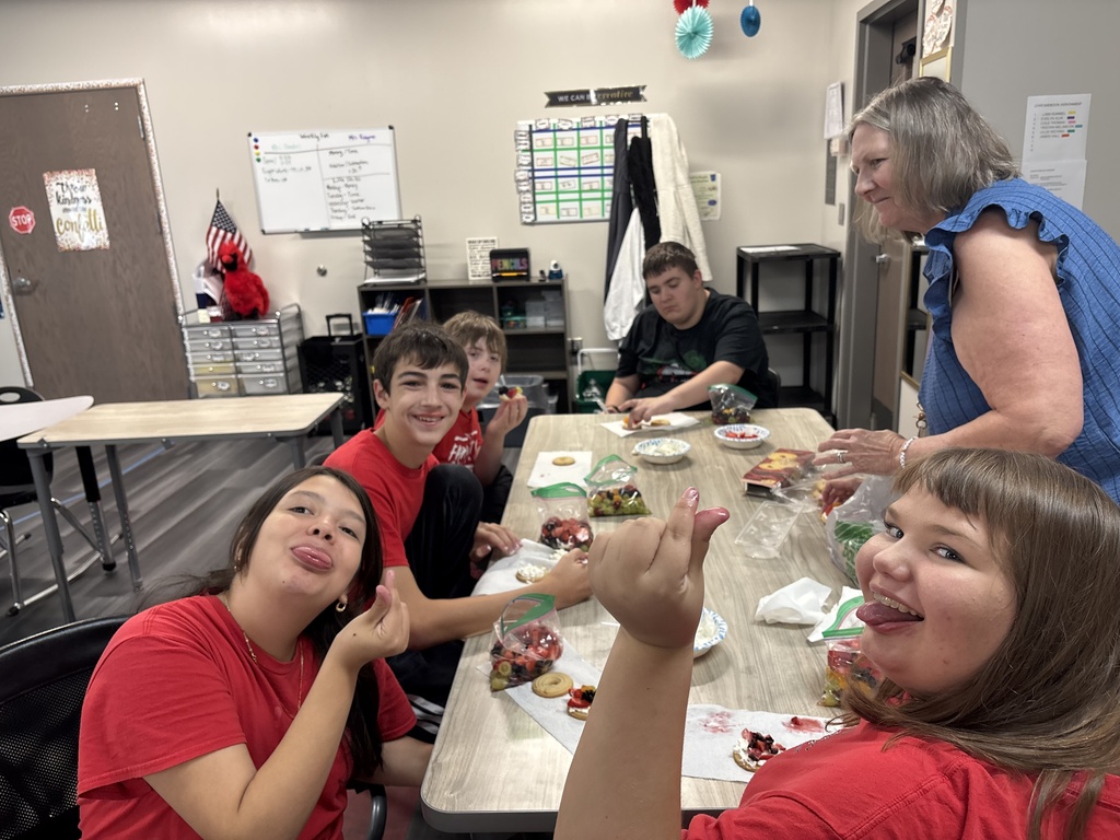 A group of students in red shirts sit around a table eating food. A woman stands beside the table.
