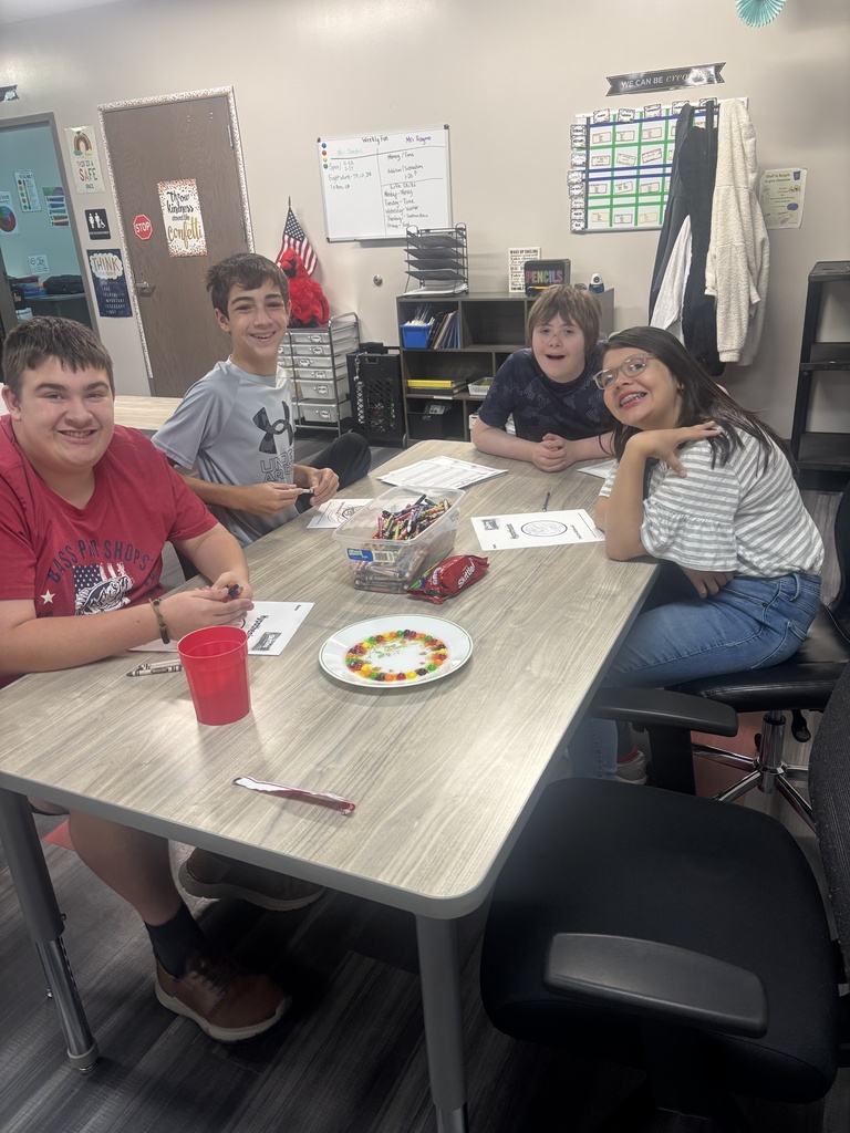Four students seated at a table with colored pencils, a red cup, and a plate with food.