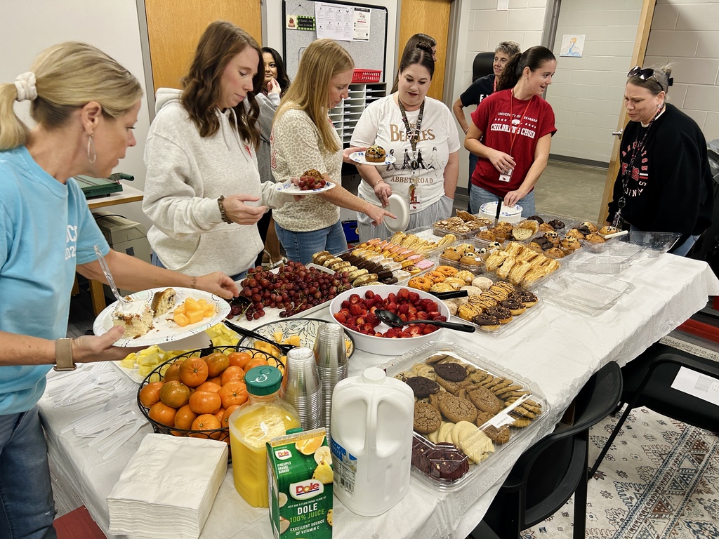 A group of women gather around a table with a variety of foods including fruit, cookies, and juice.