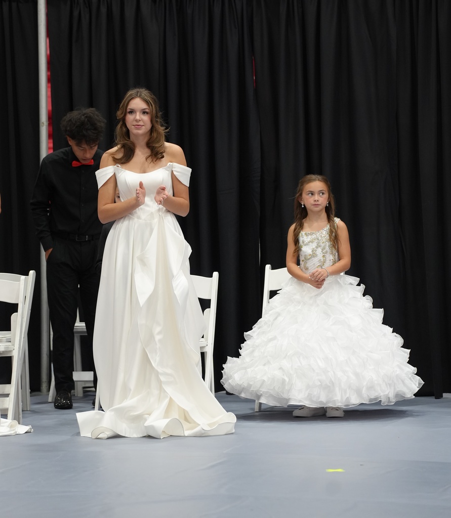 Woman in a homecoming dress stands beside a young girl in a dress on a stage with a black backdrop.