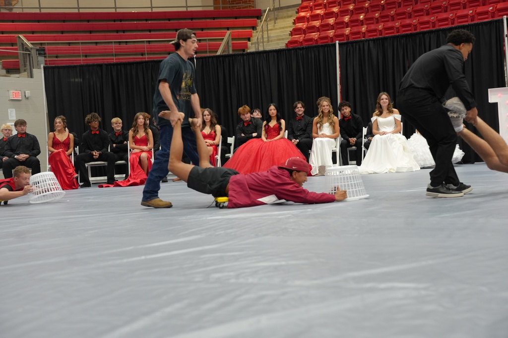 Two people perform a floor exercise in front of a crowd sitting on chairs, with red bleachers in the background.