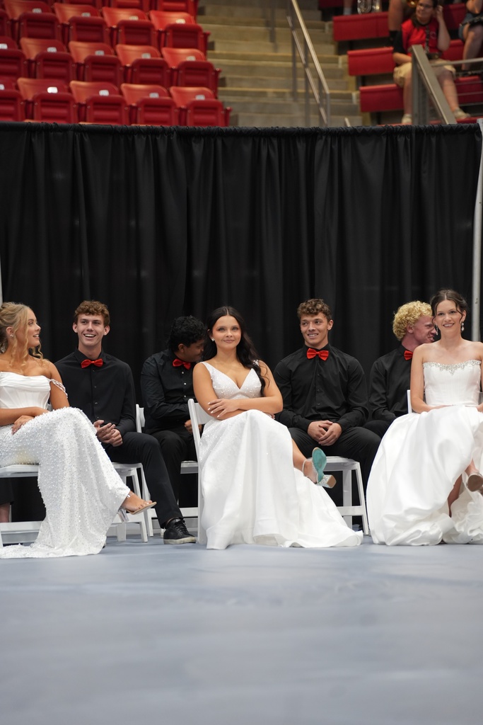 Several people in white gowns sit on chairs in front of a black curtain. Behind them are empty red chairs.