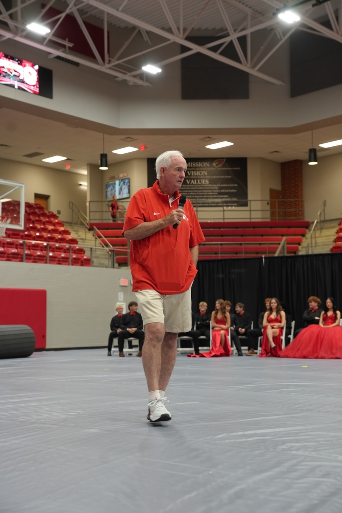 Man in orange shirt and white shorts speaking into a microphone on a stage with rows of red chairs.