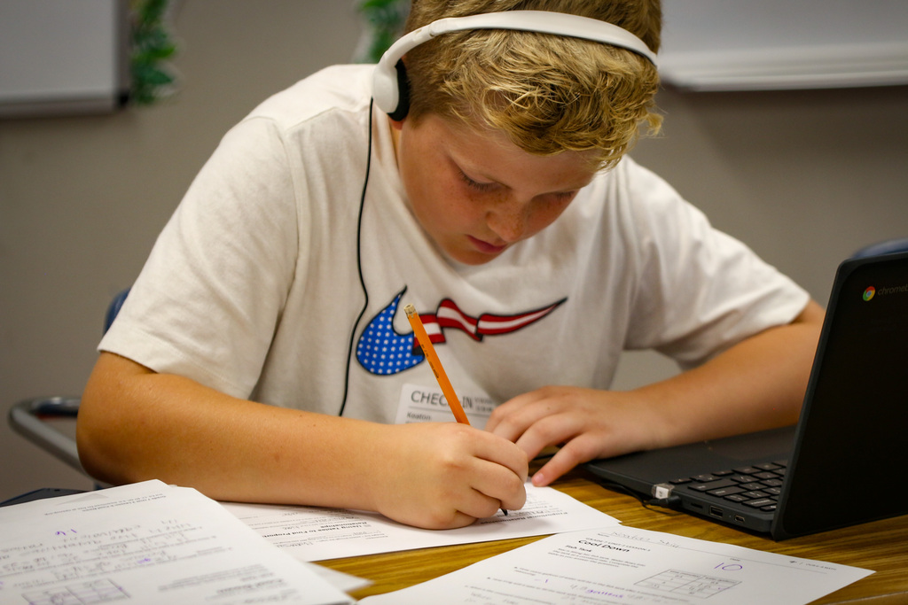 A student wearing headphones writes on paper with a pencil, focused on a laptop on the desk.