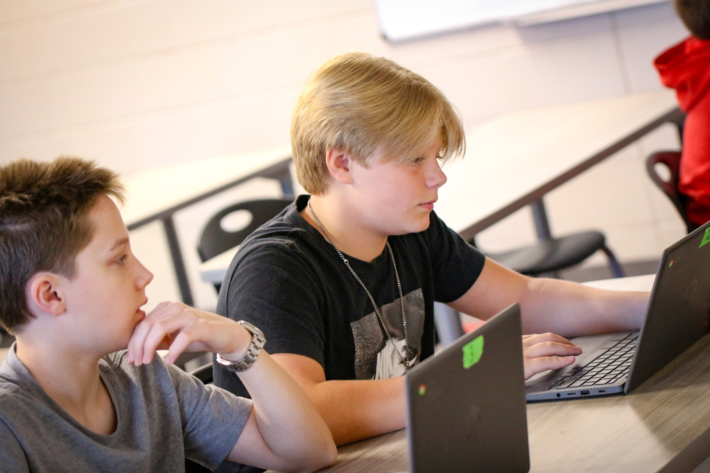Two young boys sit at a table, each working on a laptop. One has a green tag on the laptop.