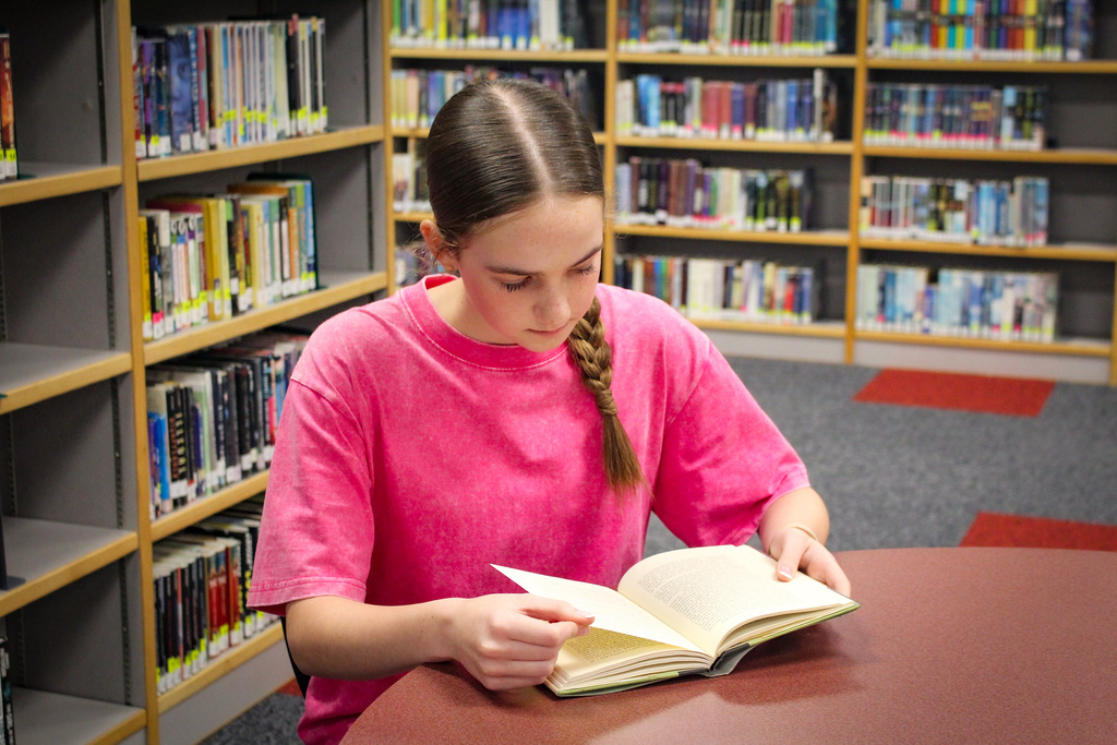 A young girl wearing a pink shirt sits at a library table, focused on an open book.