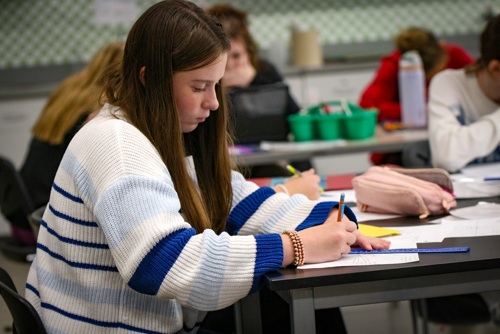 A student works on papers at a desk with others in the background in a classroom.