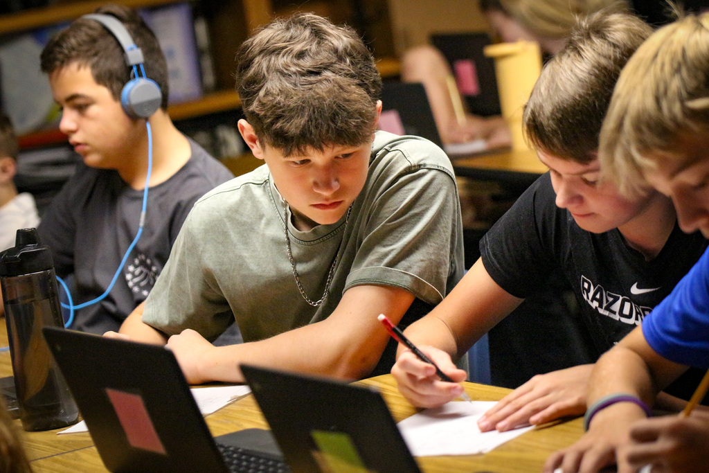 Four boys in a classroom, using laptops and writing on paper.