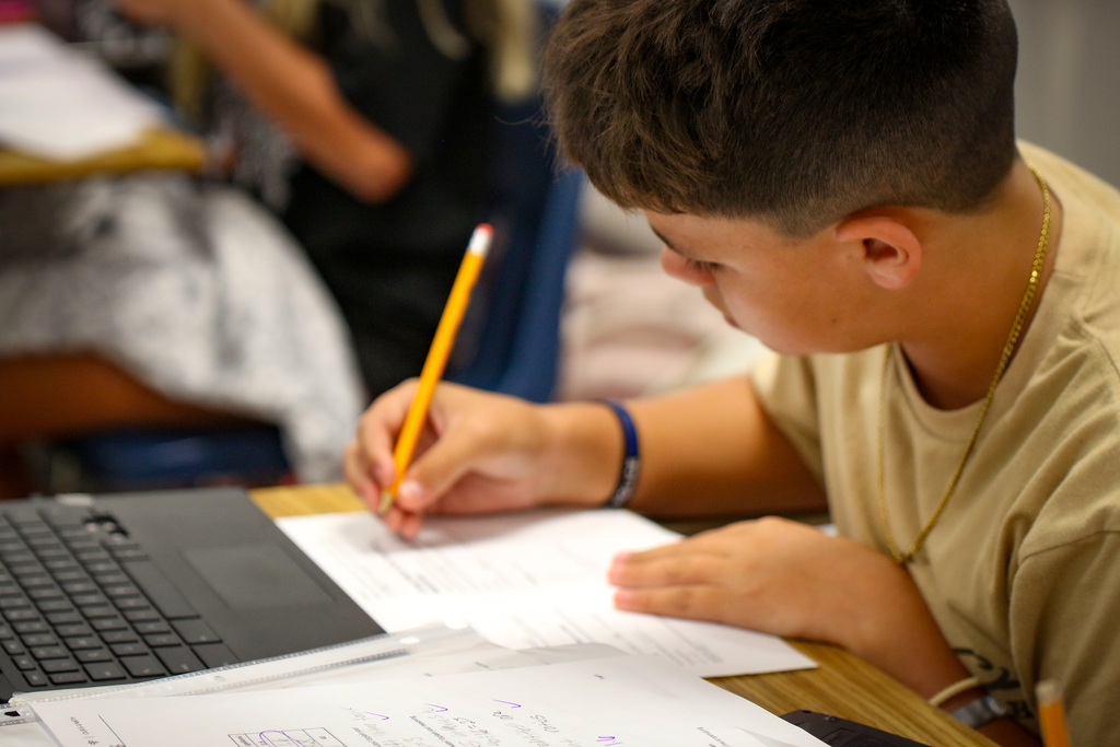 A young student in a tan shirt focuses on writing with a pencil on paper, a laptop nearby.