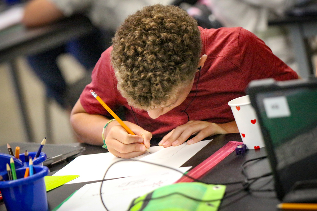 A young person draws with a pencil on a piece of paper, seated at a desk.