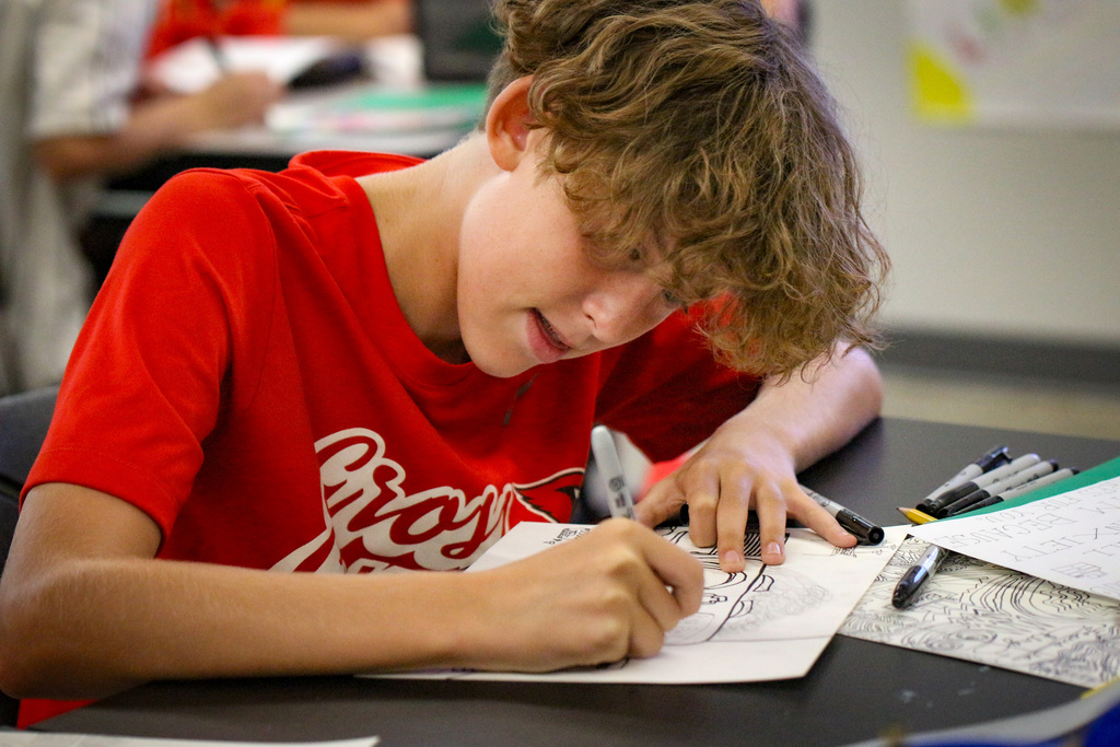 A boy in a red T-shirt draws on paper at a desk with pens and markers.