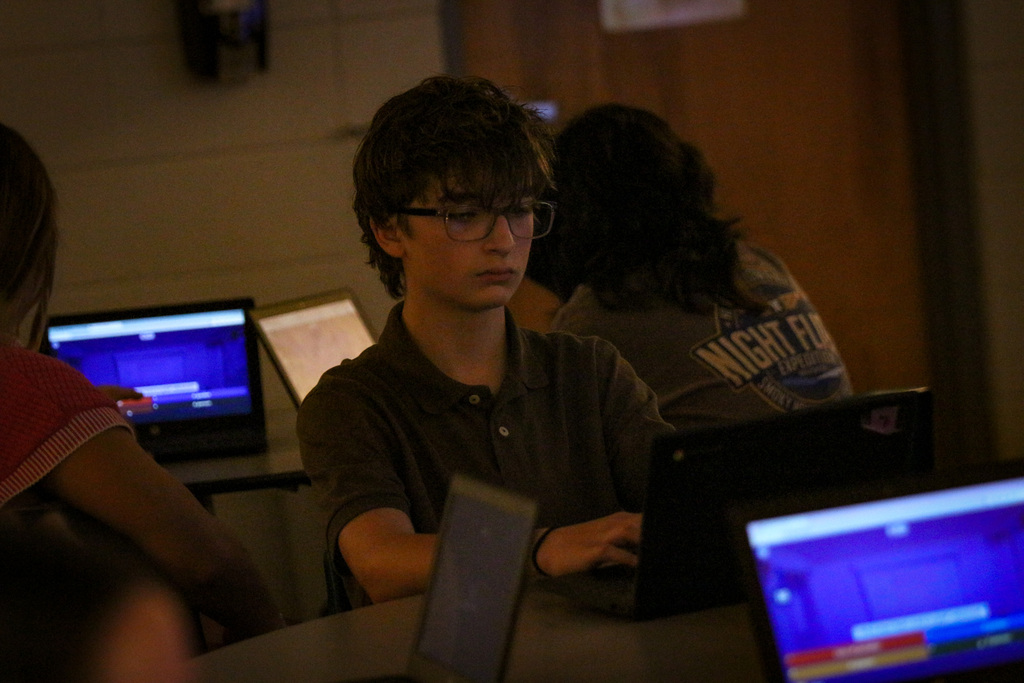 Students sit around a table using laptops. A boy wears glasses. One screen shows a blue background.