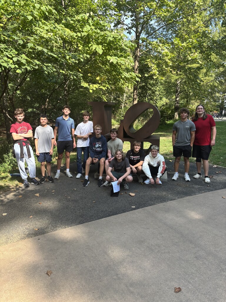 Group of individuals in various colors, including white, blue, red, and gray, posing in a park with a large letter sculpture.