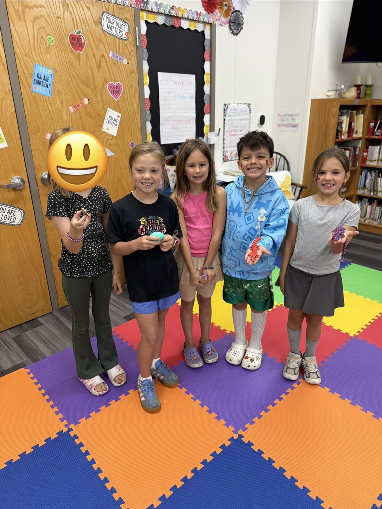 Five children stand on a colorful floor mat in a classroom, wearing casual clothes. They are smiling and posing for a photo.