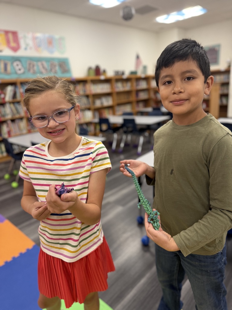 Two children hold craft items, one in a skirt and the other in a shirt, in a room with bookshelves.