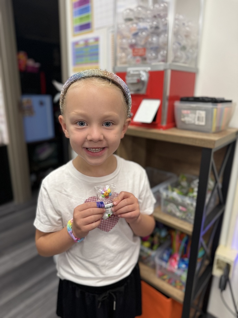 A child in a white T-shirt and black skirt holds a candy package, smiling. Behind is a shelf with assorted candies.