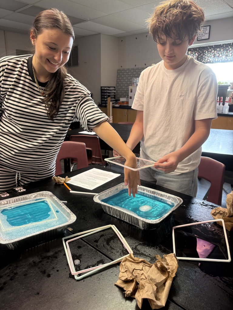 Two students work at a table with four trays of blue liquid, one holding a frame.